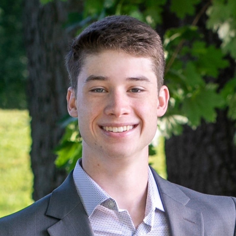 A young man with short, brown hair and fair skin is smiling at the camera. Dressed in a grey suit jacket and white collared shirt, he stands outdoors amidst green leaves and tree trunks. This photo is perfect for digital marketing services, focusing attention on his face with a beautifully blurred background.