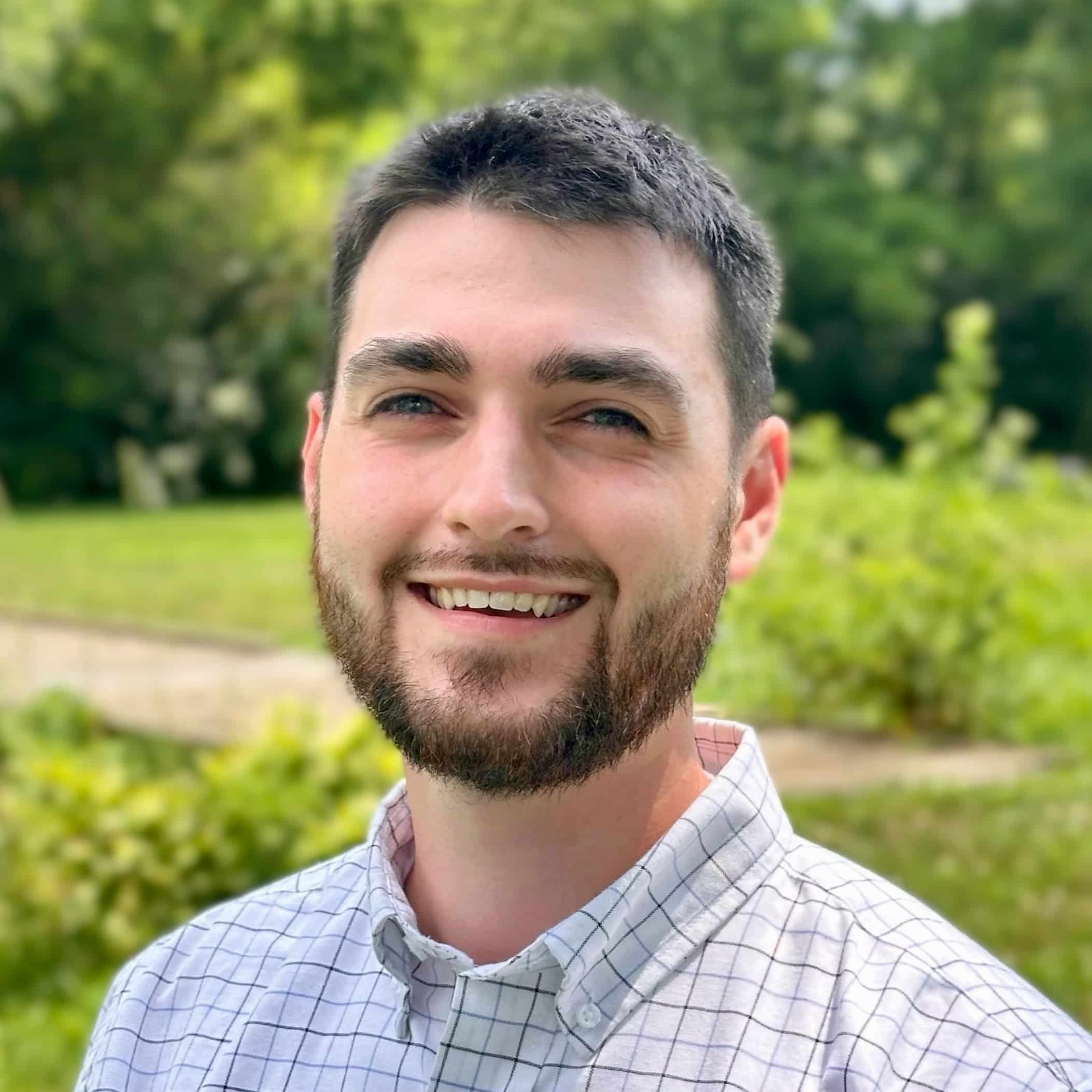 A man with short dark hair and a beard smiles at the camera. He is wearing a light-colored checkered shirt and standing outdoors in a lush, green area with trees and bushes in the background. The lighting is bright, suggesting a sunny day—perfect for an "About" page on your website.